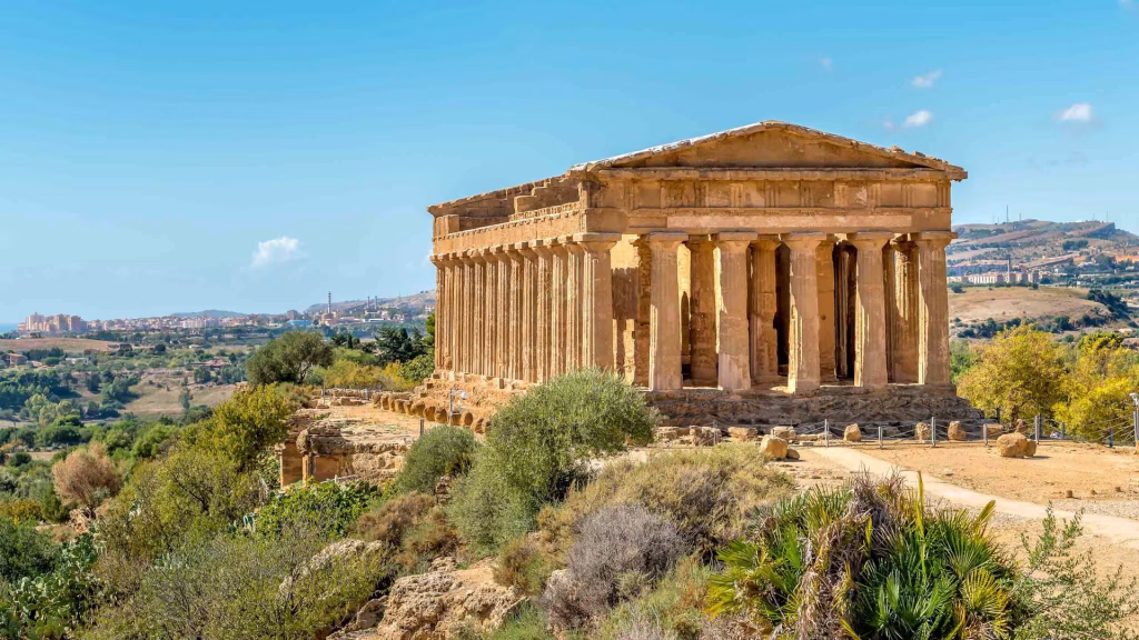 Doric temple with rows of columns on a hilltop, with fields and town below under blue sky.