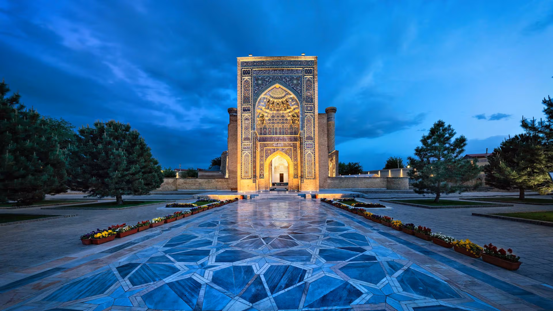Ornate tile-arched gateway centered in a paved courtyard with flower beds and a twilight blue sky behind it.