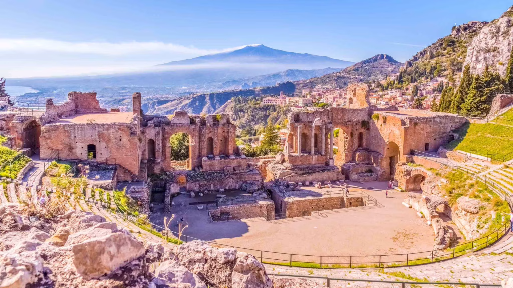 Ruined ancient Greek theatre with stone seating, Mount Etna smoking in the distance.