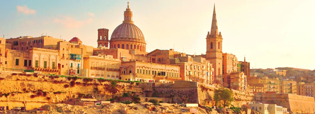 Sunlit cityscape with a large domed cathedral and a tall spire among warm stone buildings along the waterfront.
