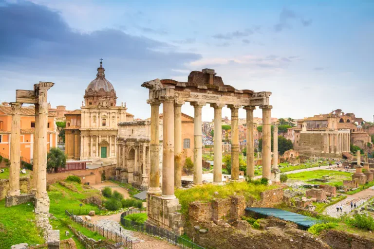 Ruins of the Roman Forum with tall ancient columns, a domed church nearby, and historical buildings in the background.