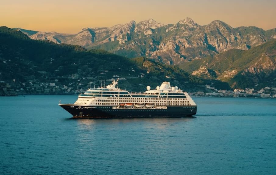 Large cruise ship sailing on calm blue water with rugged green mountains and a golden sunset in the background?