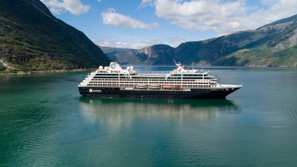 Large cruise ship sailing through a narrow fjord with steep green mountains on either side, calm blue-green water beneath.