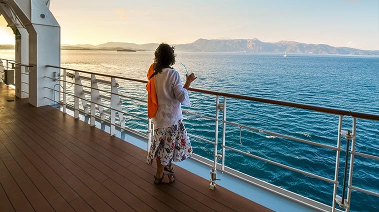 Woman standing on a cruise ship deck, looking at the sea at sunset, wearing a floral dress, white cardigan, and an orange bag across her shoulder.