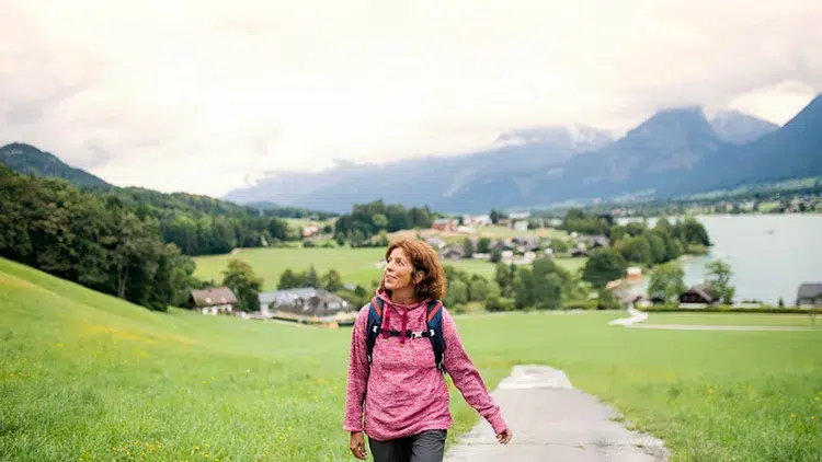 Woman wearing a pink jacket and backpack walks along a path through green countryside with a lake and distant mountains ahead.