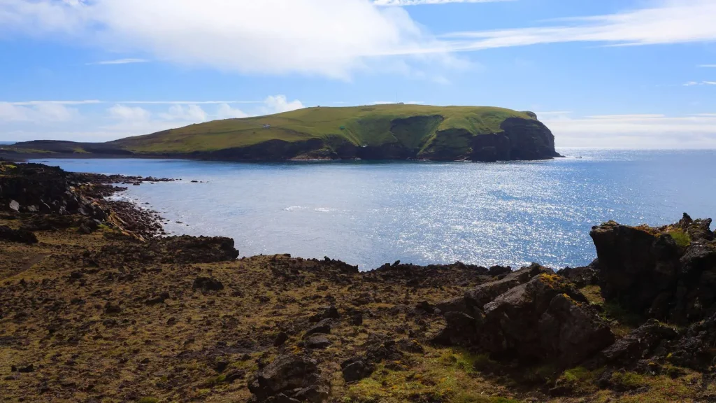 rocky shoreline in the foreground leads to a calm sea and a grassy, cliff-sided island under a blue sky