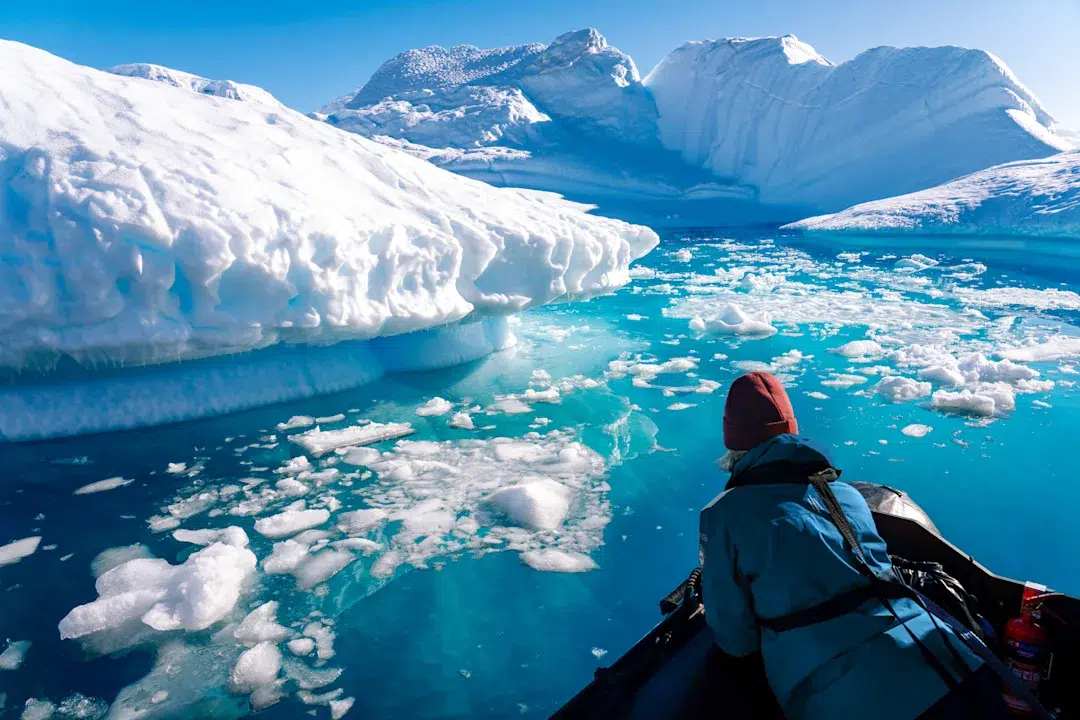 Person in a blue jacket and red hat paddles a small boat toward a towering iceberg in turquoise Arctic waters.
