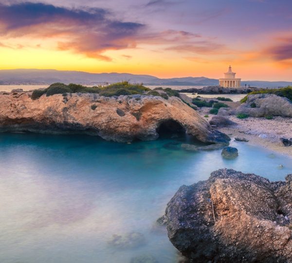 Sunset over a rocky coastline with turquoise water and a distant round temple on the shore at the horizon.