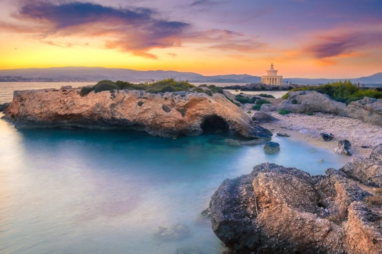 Sunset over a rocky coastline with turquoise water and a distant round temple on the shore at the horizon.