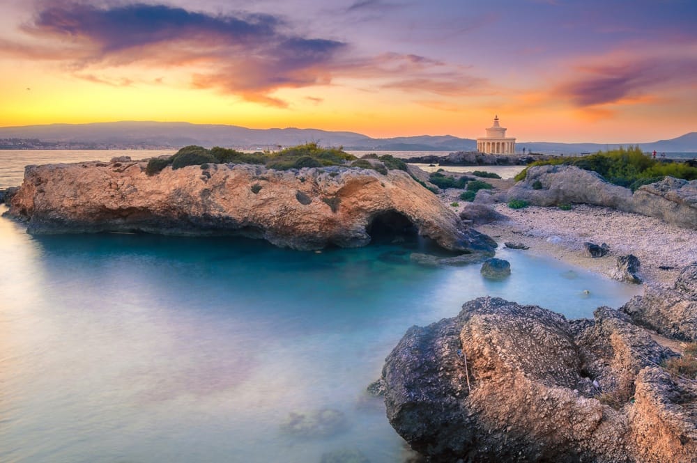 Sunset over a rocky coastline with turquoise water and a distant round temple on the shore at the horizon.