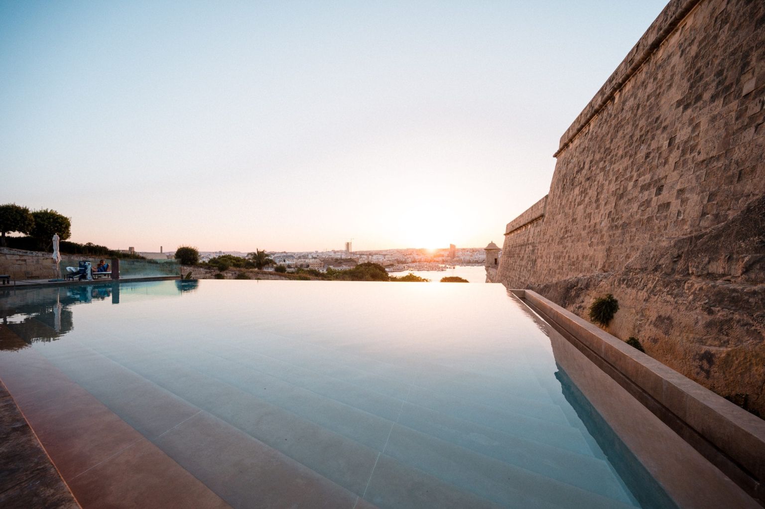 Infinity pool at sunset with a tall stone fortress wall on the right and a distant city on the horizon reflecting in the water