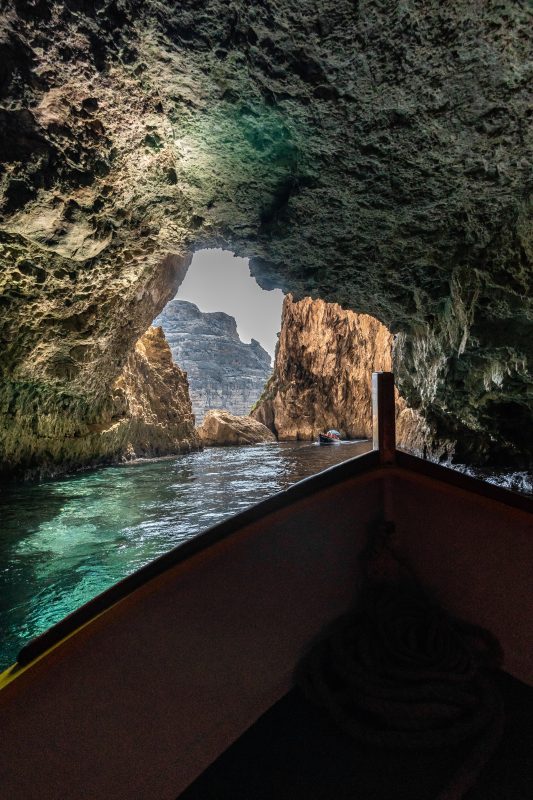 Boat rides through a rugged sea cave with green water and a bright opening in the distance.
