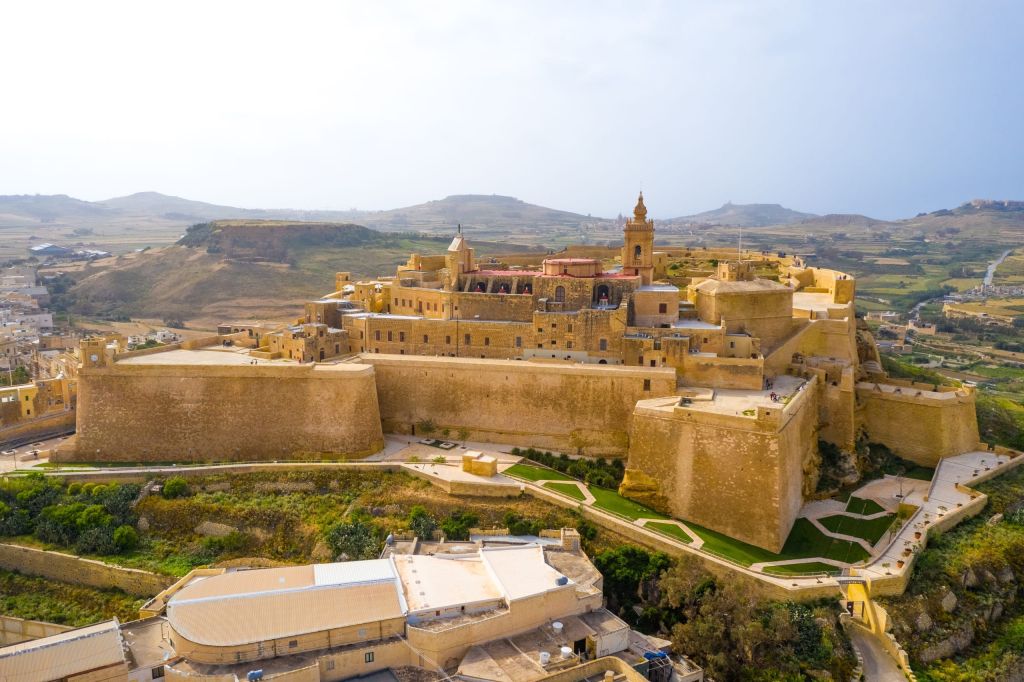 Bird's-eye view of a historic limestone fortress city with thick walls, bastions, and a tall bell tower on a hill overlooking a rural landscape.