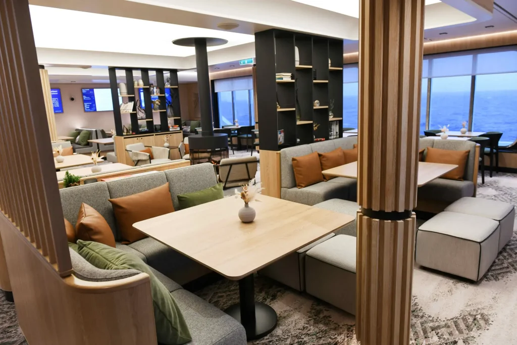 Open-concept lounge with gray sectional sofas, rust and green cushions, light wood tables, and wooden slat partitions, overlooking the sea through large windows.