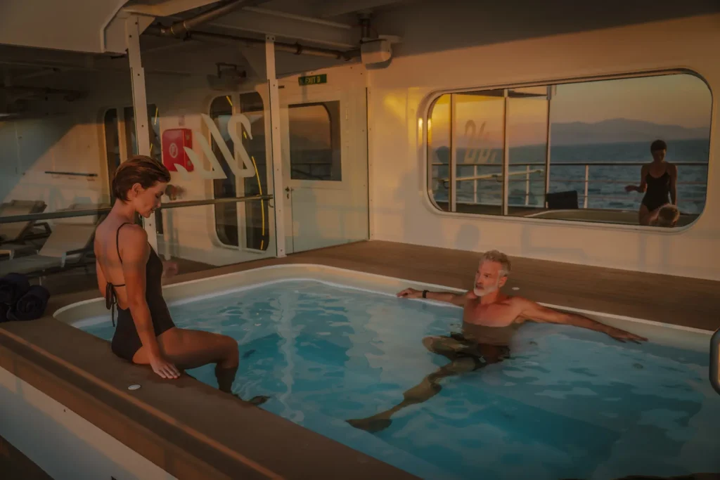 Indoor yacht pool scene with two adults in swimsuits relaxing near the edge as sunset lights the white cabin; ocean seen through large window in the background.