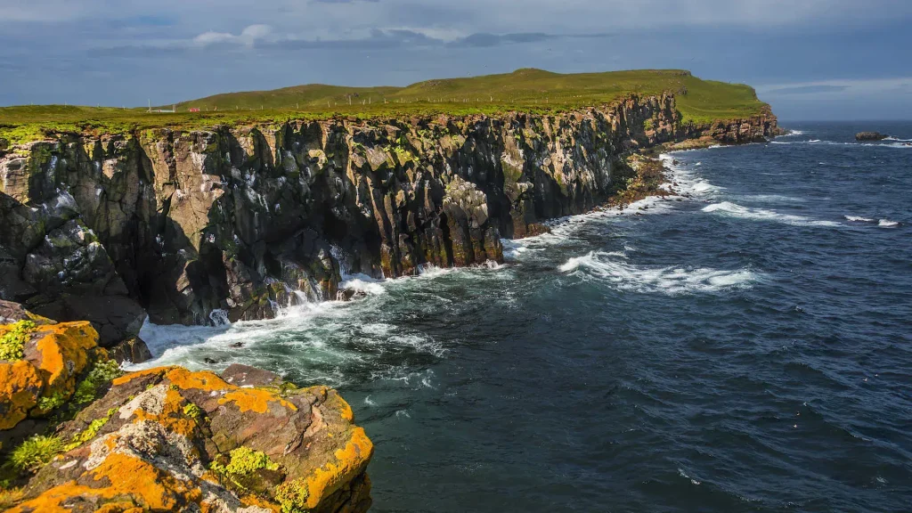 Rugged coastal cliffs with green grass atop, waves crashing against jagged rocks along a blue sea.