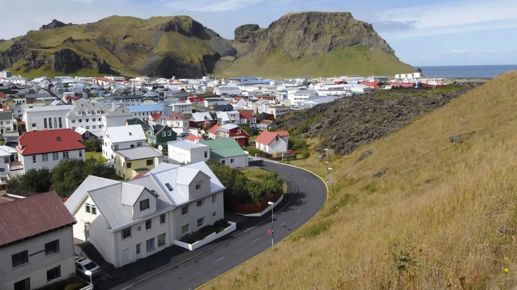 Coastal town with pastel houses along a winding road, green hills behind and the sea to the right.