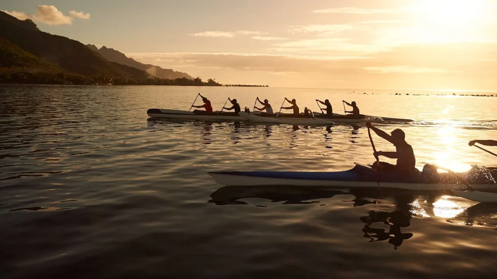 Silhouetted rowers in a multi-person canoe gliding across calm water at sunset, with mountains in the background and a golden sky.