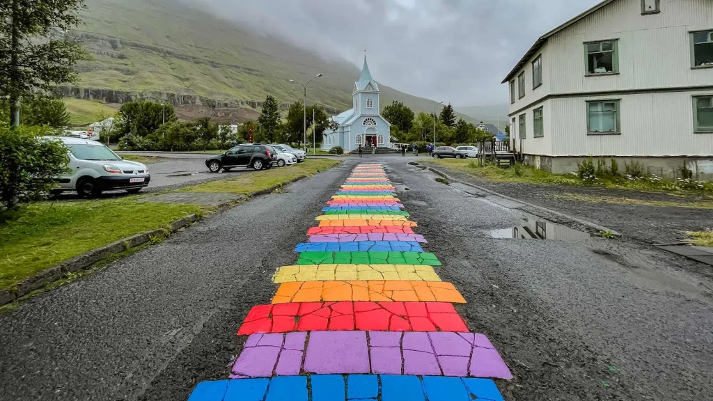 Rainbow-painted stepping stones lead to a blue church in a small town, overcast sky above mountains.