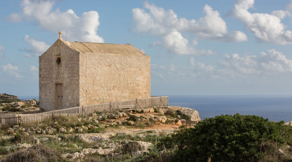 Stone chapel perched on a rocky cliff, cross on top, with the sea and blue sky in the background.