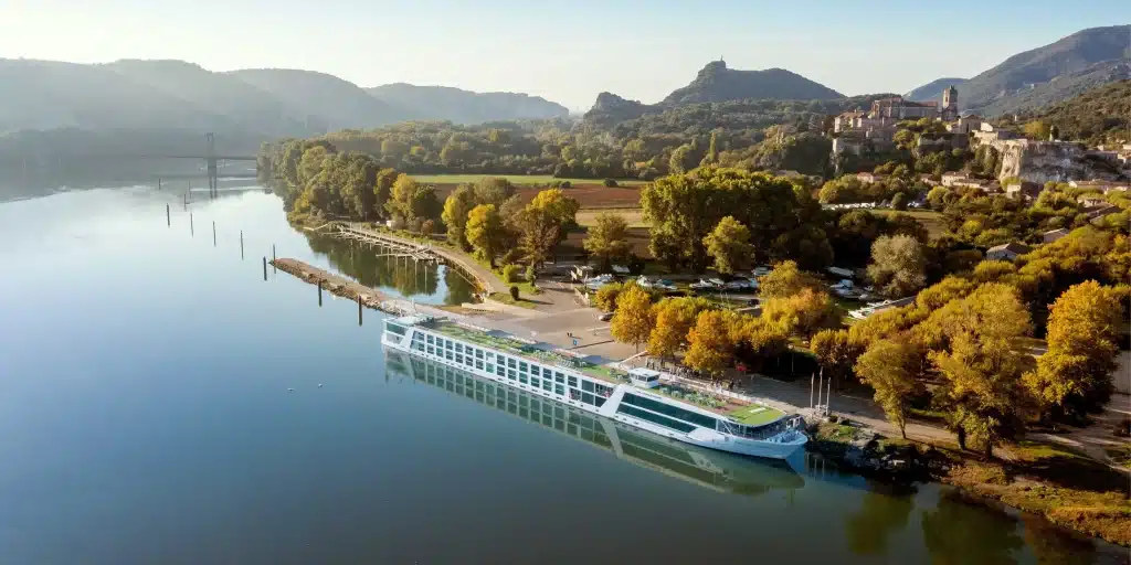 River cruise ship moored on a calm river beside autumn trees and a hilltop village.