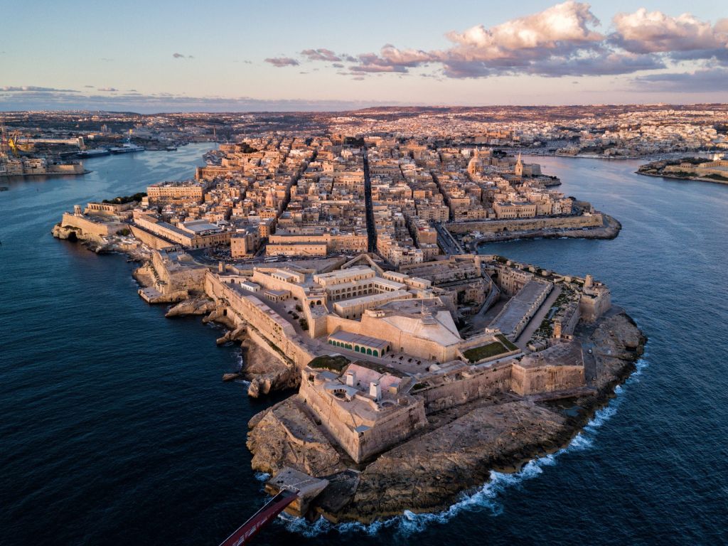 Aerial view of a fortified harbor city on a sunlit peninsula with limestone buildings and blue sea around it, likely Valletta, Malta.
