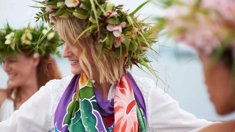 Woman with a large floral crown and colorful scarf smiling outdoors at a festival or parade.