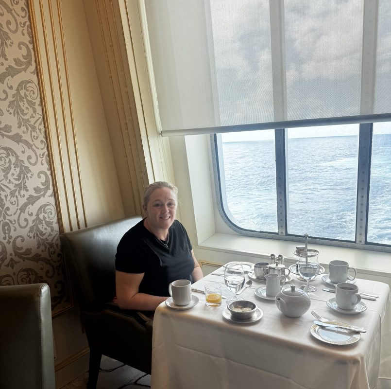 Woman seated at a formal dining table on a ship, with a white tablecloth and tea set, overlooking the ocean through a large window.