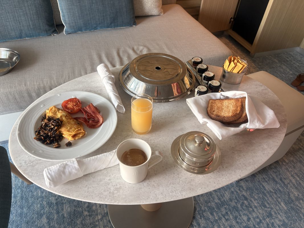Plate with breakfast: omelette, beans, bacon, sliced tomato and peppers on the left; toast in a napkin on the right; glass of orange juice in the center; coffee cup nearby, in a cozy hotel room setting.