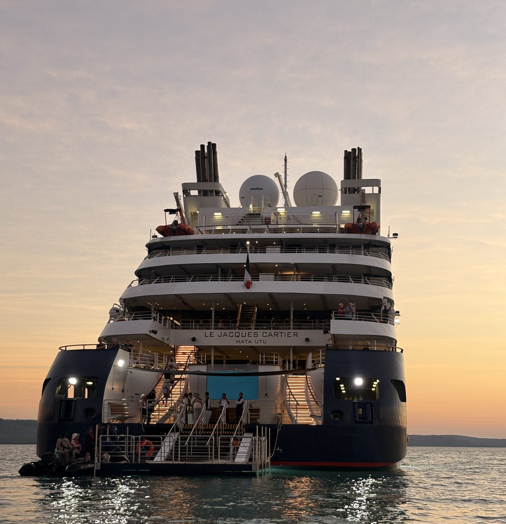 Rear view of the Le Jacques Cartier cruise ship at sunset, passengers on the decks and the ship’s name visible.