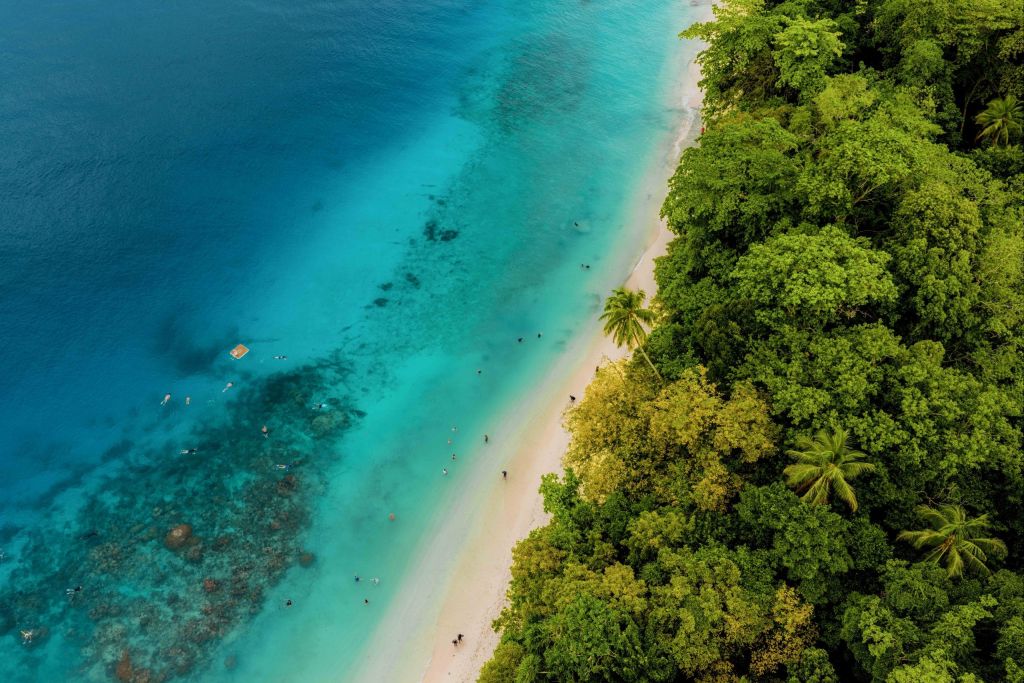 Aerial view of a tropical beach with turquoise water, white sand, and dense green jungle on the right side.
