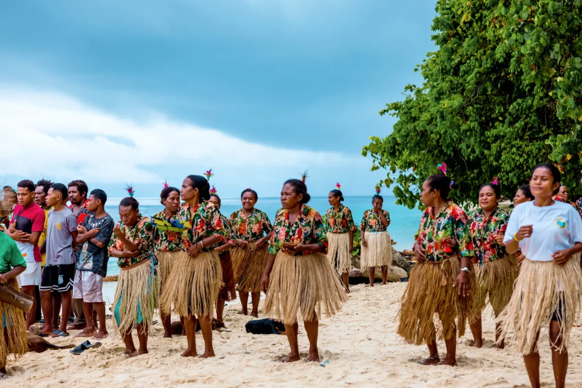 Group of people in colorful matching outfits and grass skirts performing a dance on a sandy beach, with the ocean and trees in the background.