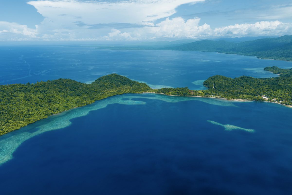 Aerial view of blue ocean with green tropical islands and white-sand beaches along a curved coastline. Raja Ampat cruise