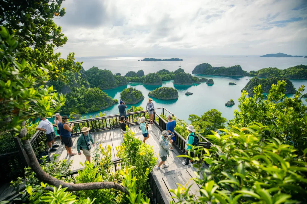 Tourists on a wooden viewpoint overlooking turquoise water and small green islets surrounded by lush jungle.