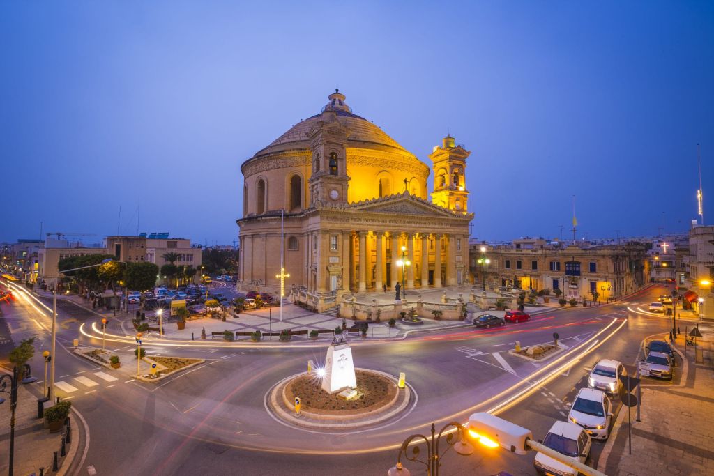 Night view of a grand domed church illuminated in warm yellow light, centered on a roundabout with streaks of car headlights, surrounded by buildings at dusk (Maltese city).