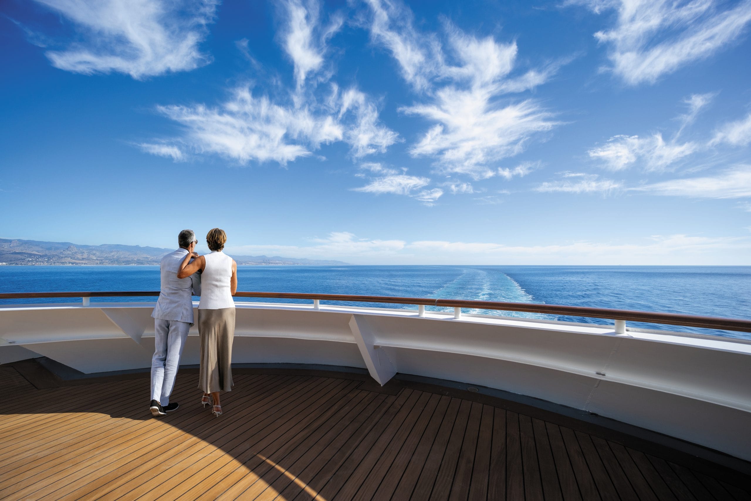 An older couple stands with their backs to the camera on a cruise ship deck, looking out over the ocean toward distant mountains under a blue sky with wispy clouds, arms around each other.