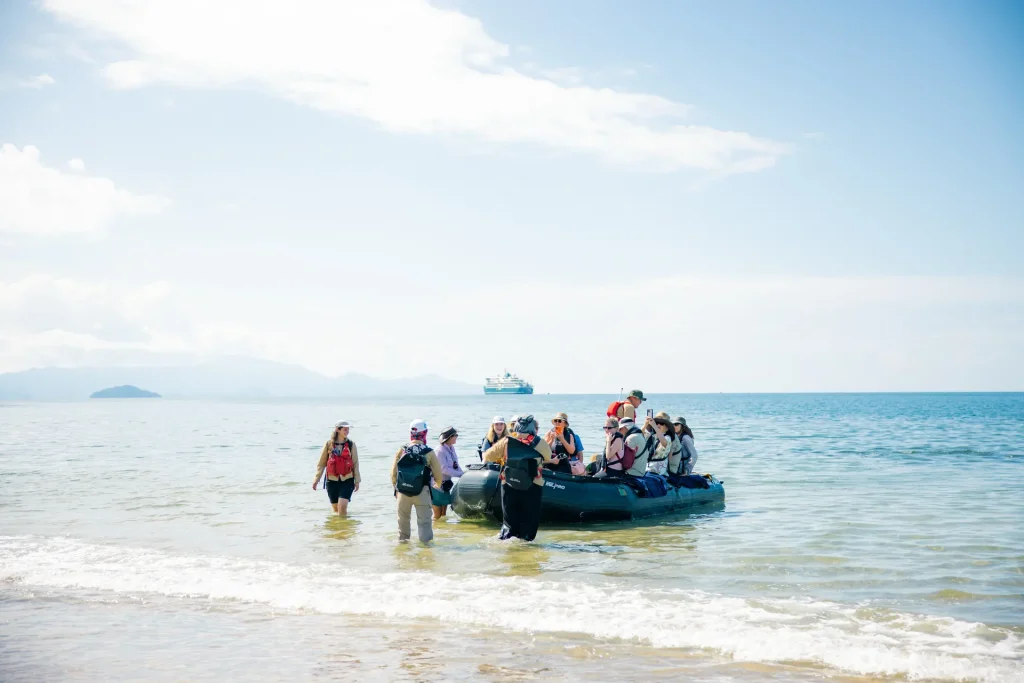 Group of people wearing life jackets gather around a small inflatable boat in shallow sea water at the beach.