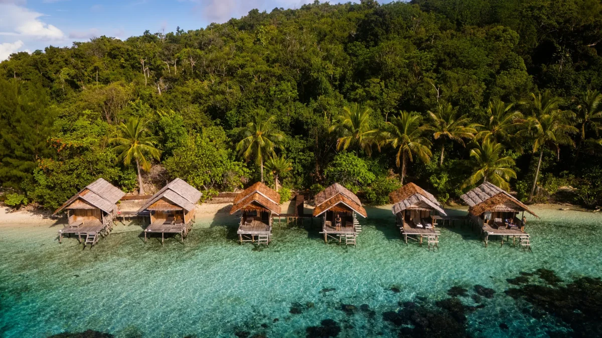 Overwater bungalows with thatched roofs extend into turquoise lagoon, backed by lush tropical forest. (informative)