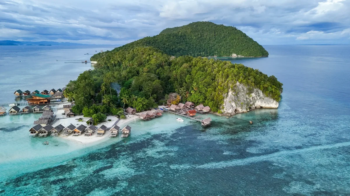 A tropical island resort with overwater bungalows along a white-sand beach and turquoise shallow water.