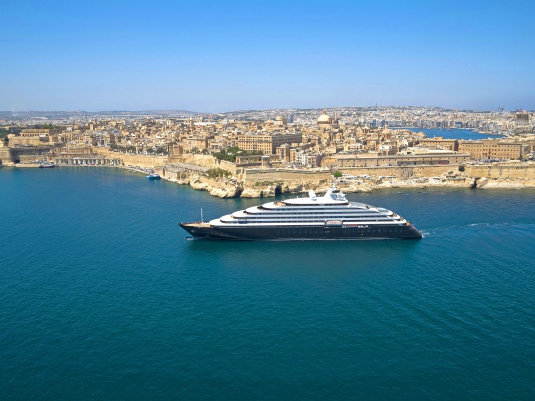 A large white multi-deck cruise ship sails near a historic limestone city along a blue harbor shoreline.