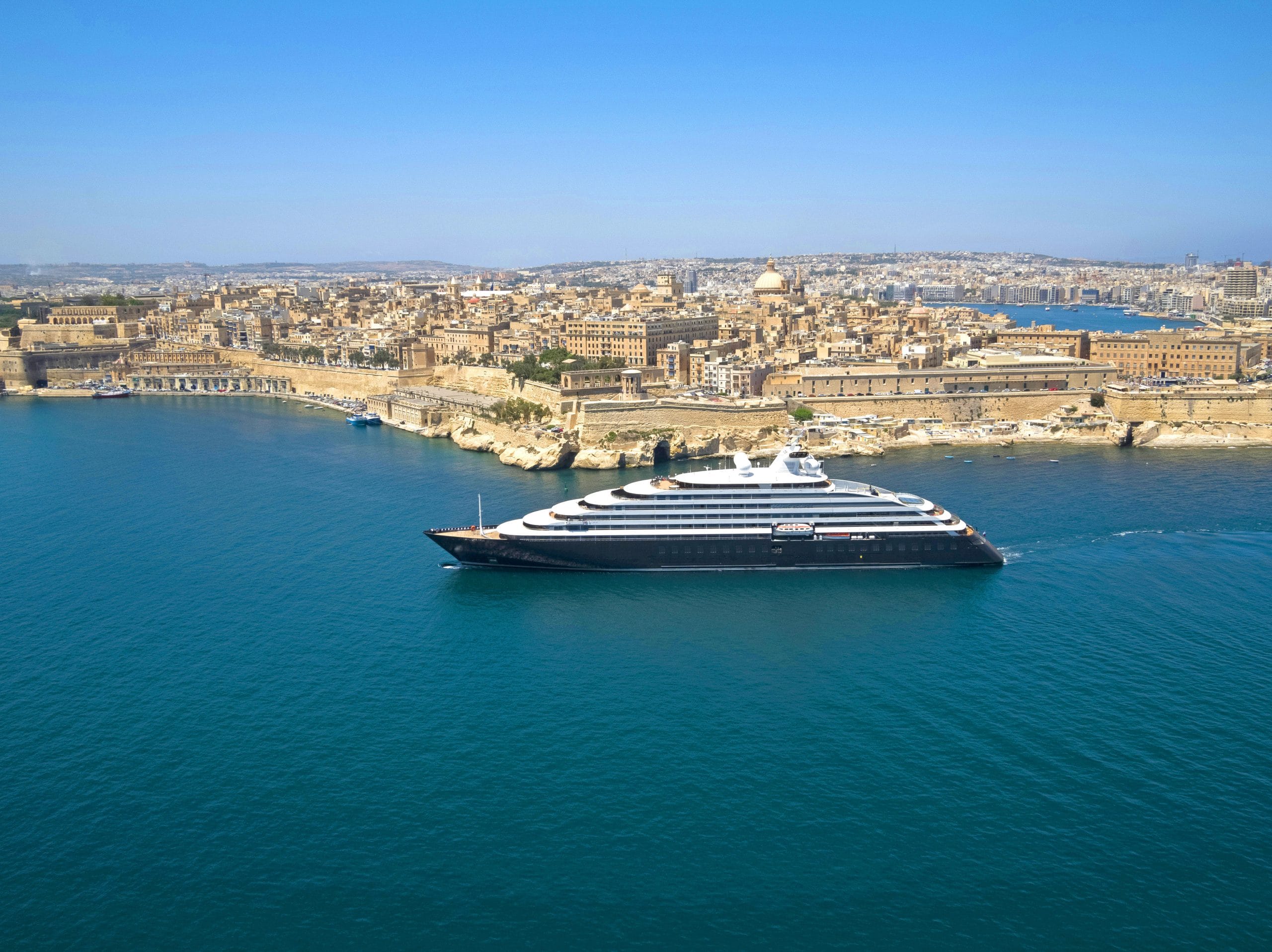 A large white multi-deck cruise ship sails near a historic limestone city along a blue harbor shoreline.