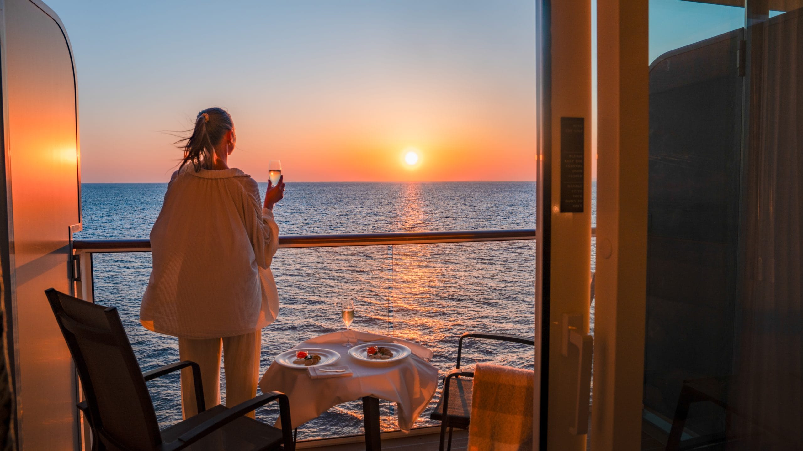 Woman on a cruise ship balcony holding a glass of wine at sunset, ocean view in the background, with a table of desserts nearby.