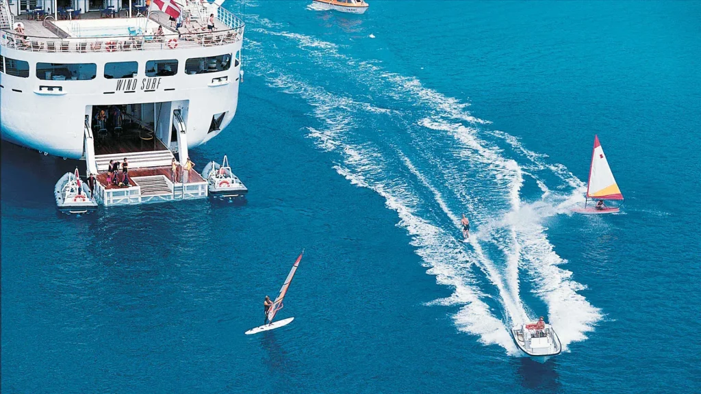 Large white cruise ship named Wind Surf at sea, with passengers on the upper deck and small support boats nearby as windsurfers ride nearby wake in bright blue water.