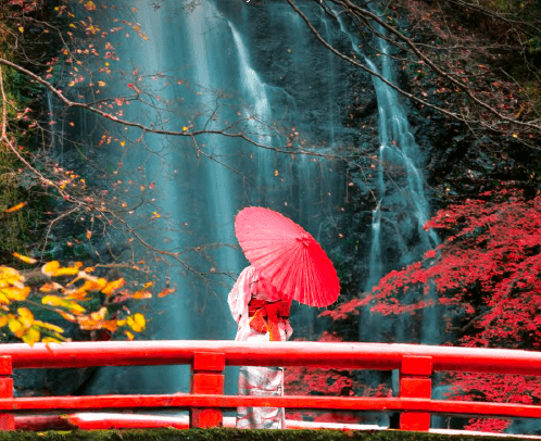 Person in traditional dress with a red umbrella standing on a red bridge beside a waterfall and autumn-colored trees, Seabourn branding above.
