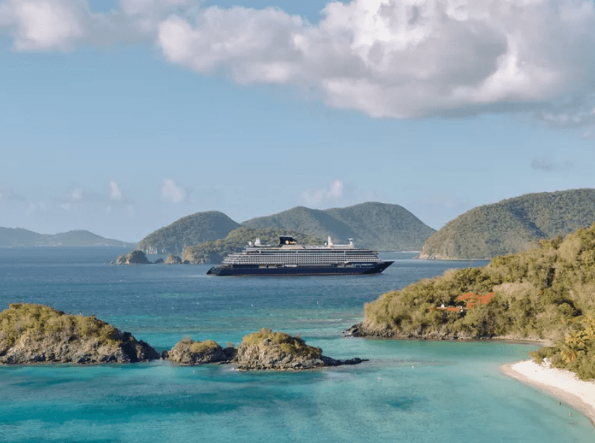 Cruise ship anchored in turquoise tropical waters with green hills and small rocky islets in the background.