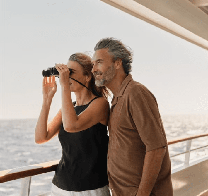 Couple on a yacht smiling at the sea, with the woman looking through binoculars from the railing.