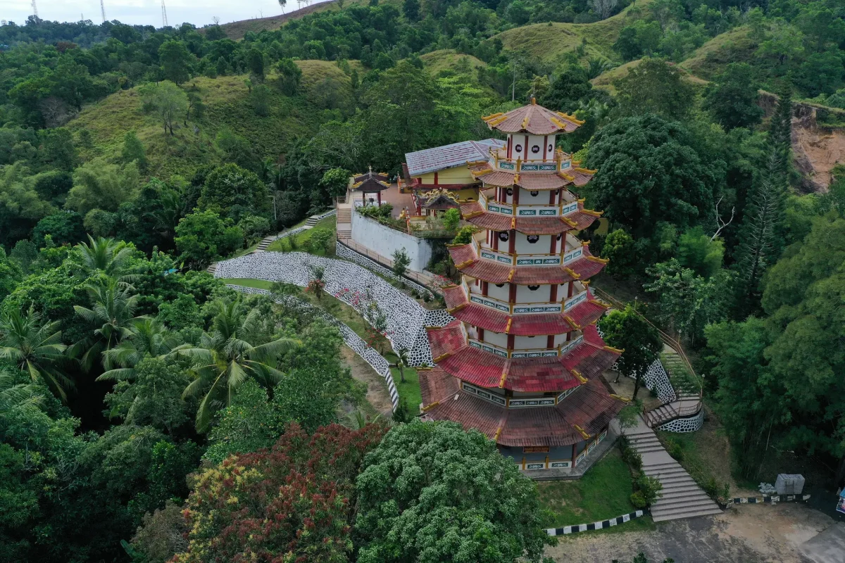 Aerial view of a multi-tiered red-roofed pagoda nestled among dense tropical trees on a hillside, with stone paths and terraces surrounding it.