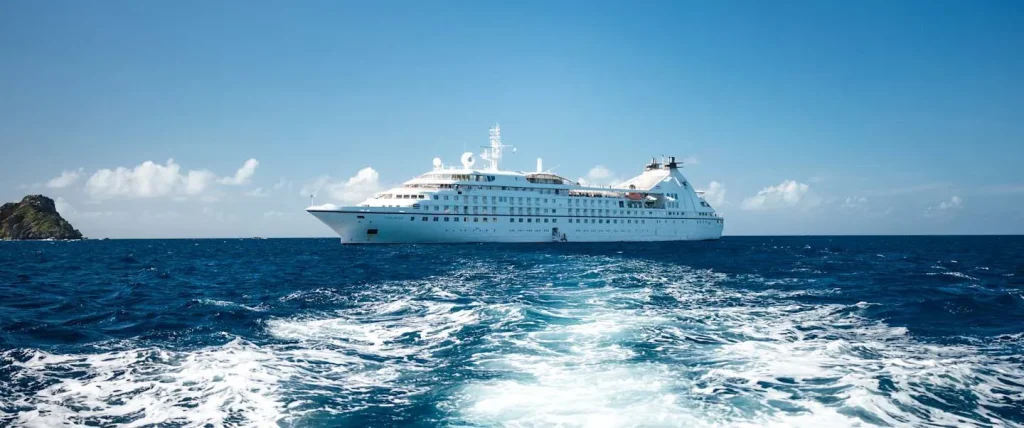 White cruise ship sailing on deep blue sea with wake trailing behind the bow area visible beneath a clear sky, left horizon shows a rocky island nearby