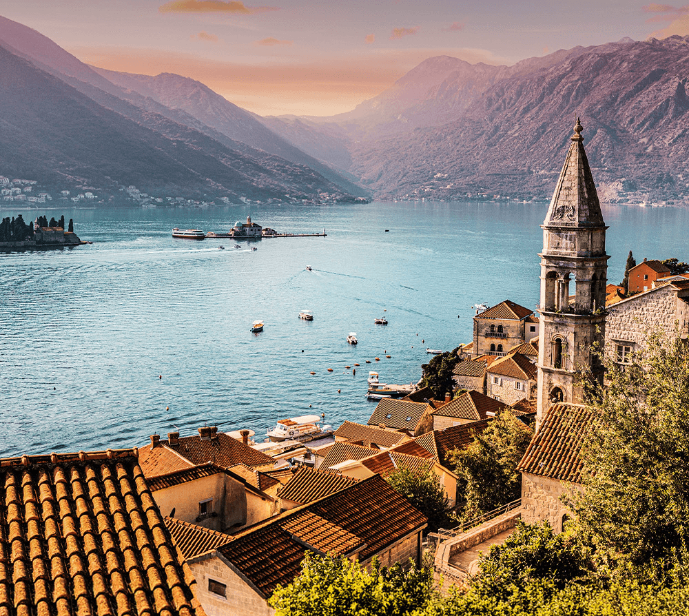 Sunset over a coastal town with red-tiled roofs and a tall stone church tower, boats on a calm blue bay, with mountains in the background.