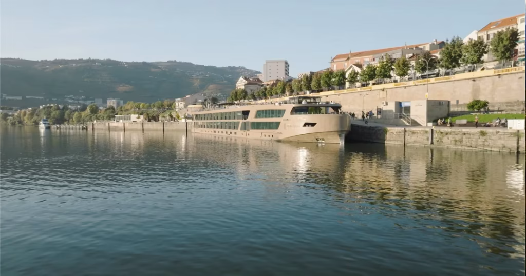 Beige river cruise ship moored at a waterfront promenade with trees and hills in the background.